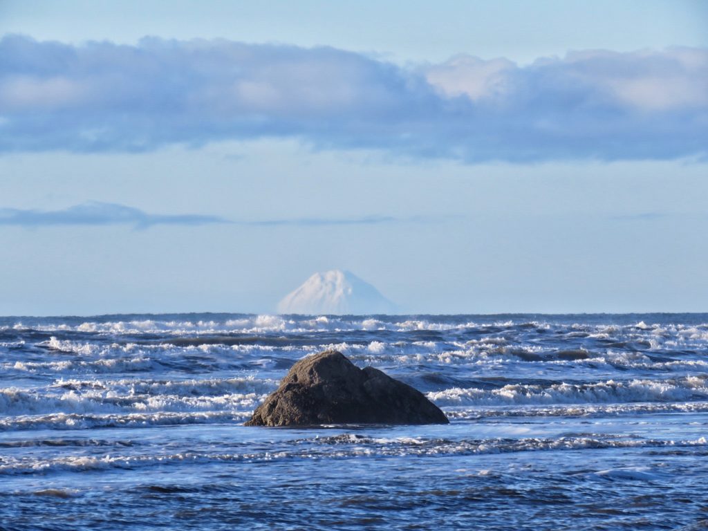 A dark stone jutting out of rough water with a snowcapped mountain behind